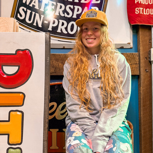 Person wearing a yellow cap and gray hoodie sitting in front of vintage signs.